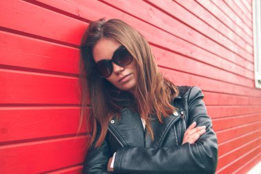 Duvarın yanında deri ceketli, kot pantolonlu ve spor ayakkabılı genç bir kadının portresi duruyor..