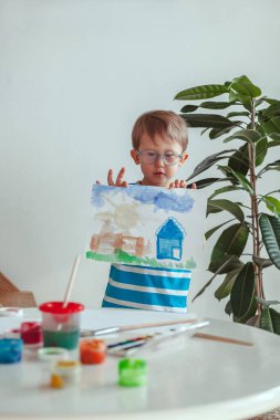 Little child boy paints with watercolors while sitting at the table, at home