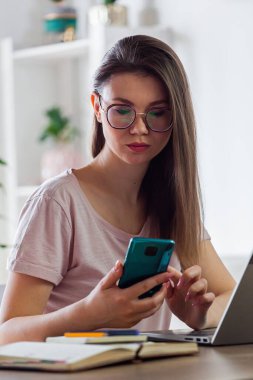 Evde çalışmak için cep telefonu ve dizüstü bilgisayar kullanan genç bir kadın.