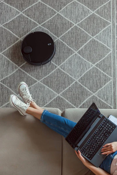 Robot vacuum cleaner cleaning carpet, woman using laptop sitting on the sofa at home