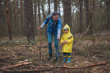 Anne ve çocuk yağmurdan sonra ormanda yürüyor. Ellerinde tahta sopalarla yağmurluklarla.