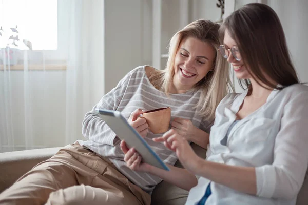stock image Two young women friends using tablet together, sitting on the couch, smiling and fun