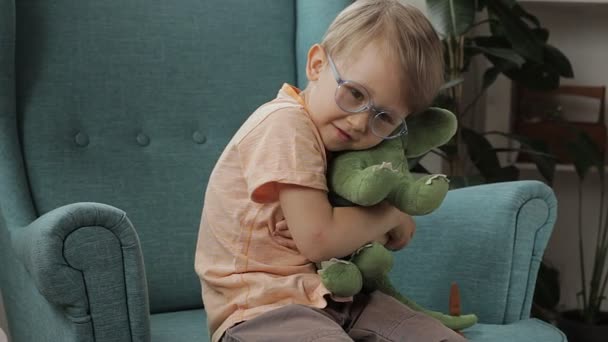 Gelukkig kind draagt bril knuffels zacht dinosaurus speelgoed zitten in fauteuil thuis — Stockvideo