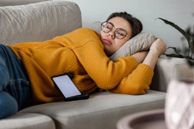 Genç bir kadın cep telefonuyla kanepede uyuyor.