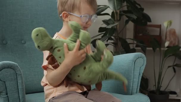 Gözlüklü mutlu çocuk evde yumuşak dinozor oyuncağına sarılıyor, okşuyor, pişman oluyor ve öpüyor. — Stok video