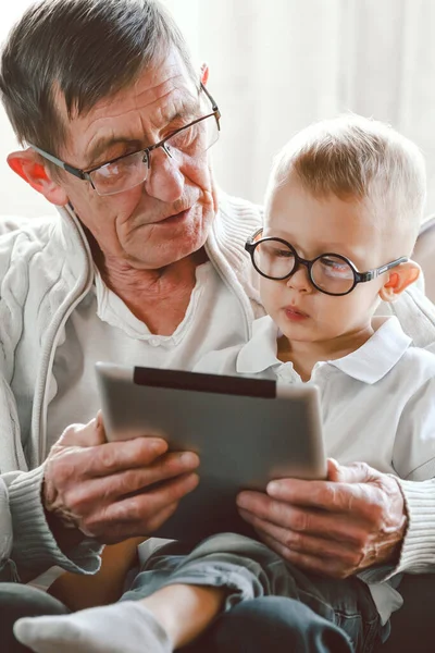 一位年迈的祖父和他的小孙子正在一起使用平板电脑 他们笑着 玩得开心 或者坐在椅子上看一段有趣的视频 — 图库照片