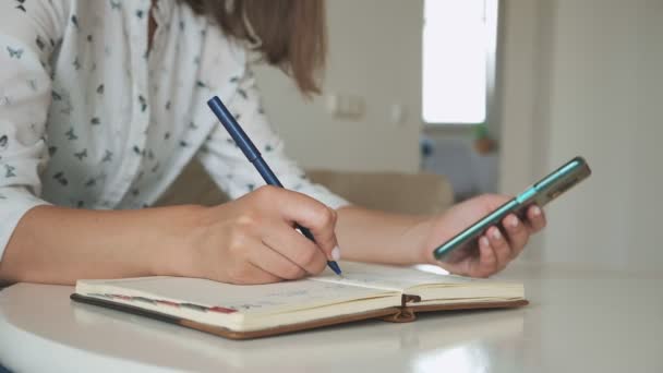 Mâini de femeie folosind telefonul mobil și scriind în notebook — Videoclip de stoc