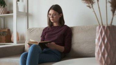 Kitap okuyan kadın evdeki kanepede oturuyor.