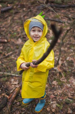 Sarı yağmurluklu çocuk yağmurdan sonra ormanda yürüyor ve elinde tahta bir çubuk tutuyor, kameraya bak.