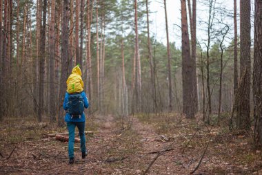 Anne ve çocuk yağmurluklarla birlikte ormanda yürüyorlar, çocuk annesinin omuzlarında oturuyor, arkadan bakıyor.