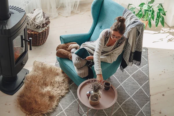 Şöminenin yanındaki genç kadın, rahat bir koltukta oturuyor, sıcak bir battaniyeyle, tablet kullanıyor.