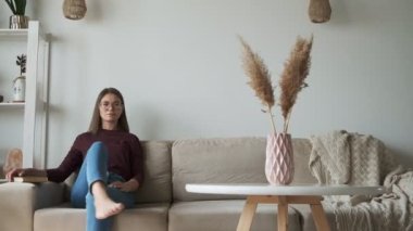 Kitap okuyan kadın evdeki kanepede oturuyor.