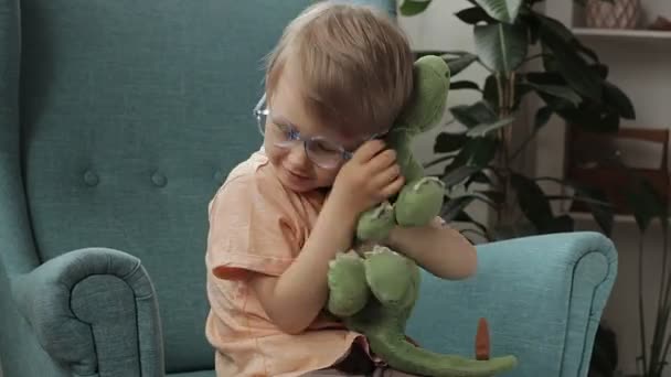 Ευτυχισμένο παιδί φορώντας γυαλιά αγκαλιάζει μαλακό παιχνίδι δεινόσαυρος κάθεται στην πολυθρόνα στο σπίτι — Αρχείο Βίντεο