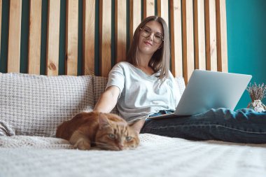 Genç bir kadın çalışmak için dizüstü bilgisayar kullanıyor, yatak odasında, evde evcil bir kediyle yatakta yatıyor. Esnek saatler ve uzak çalışma