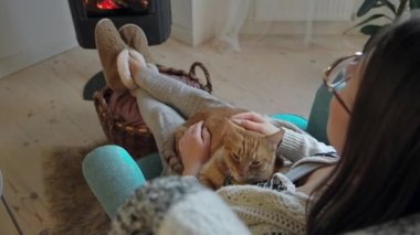 Genç bir kadın, rahat bir şekilde şöminenin başında oturmuş, kızıl bir kediyi okşuyor..