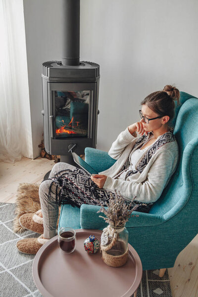 Young woman by the fireplace, sitting in a cozy armchair, with a warm blanket, using a tablet