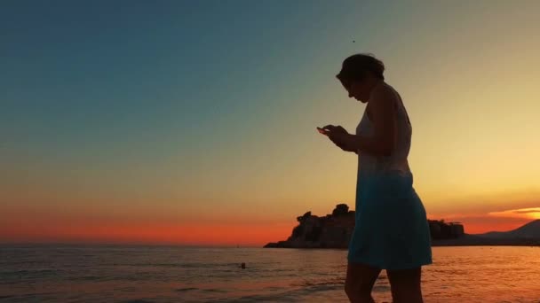 Девушка ищет в смартфоне на пляже заката — стоковое видео