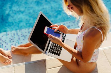 Kadın plaj havuzunda dizüstü bilgisayar ve akıllı telefon kullanıyor.