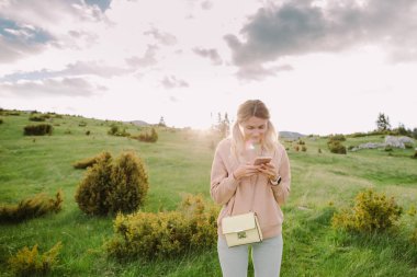Akıllı telefon kullanan mutlu kız gün batımında yeşil çayırda dinlenir.