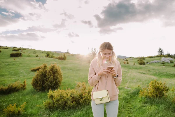 สาวที่มีความสุขที่ใช้สมาร์ทโฟน ผ่อนคลายที่ทุ่งหญ้าสีเขียวในชนบทตอนพระอาทิตย์ตก — ภาพถ่ายสต็อก