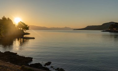 Güneş deniz ufkunun hemen üstünde kayalık plajların panoramasıyla bir gündoğumu / günbatımı yaratıyor.