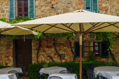 Kafe terası, krem rengi şemsiyeler, taş duvar, tırmanma asmaları, yeşil kepenkler, sıcak Toskana seti..