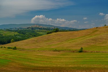 Güneşli Toskana tepeleri, yemyeşil tarlalar, uzak köy, dağınık bulutlar, sakin kırsal manzara..