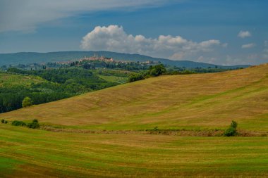 Güneşli Toskana tepeleri, yemyeşil tarlalar, uzak köy, dağınık bulutlar, sakin kırsal manzara..
