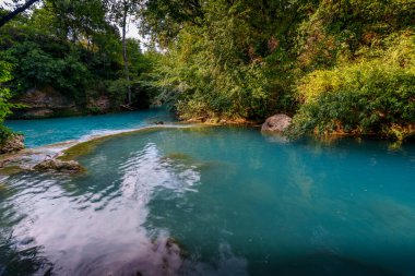 Sakin turkuaz nehir havuzu, parlak yeşil ormanlarla çevrili, huzurlu bir yaz gününde güneş ışığını yansıtıyor..