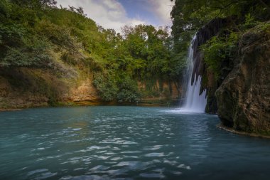 Görkemli şelale, kayalık uçurumlar ve yoğun yeşil ormanlarla çevrili derin turkuaz havuzuna dökülüyor..