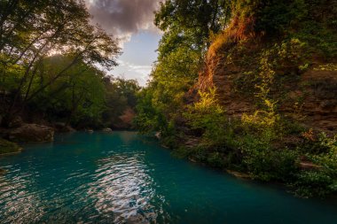 Turkuaz nehir, güneşli kayalık kayalıkların yanında esiyor. Gün batımında orman yeşilliklerini ve akşam gökyüzünü yansıtıyor..