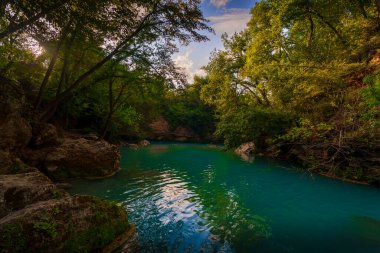 Turkuaz nehir, güneşli kayalık kayalıkların yanında esiyor. Gün batımında orman yeşilliklerini ve akşam gökyüzünü yansıtıyor..