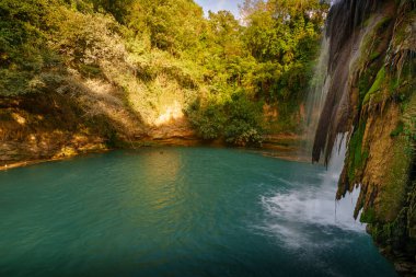 Şelalenin altındaki turkuaz havuz, yosunlu kaya yüzeyi ve ılık yaz akşamında yoğun yeşil orman..