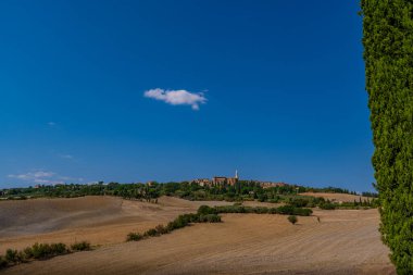 Pienza kasabası altın tarlaların ve açık Toskana gökyüzünün altındaki yeşilliklerin üzerindeki bir tepenin üzerinde görünür..