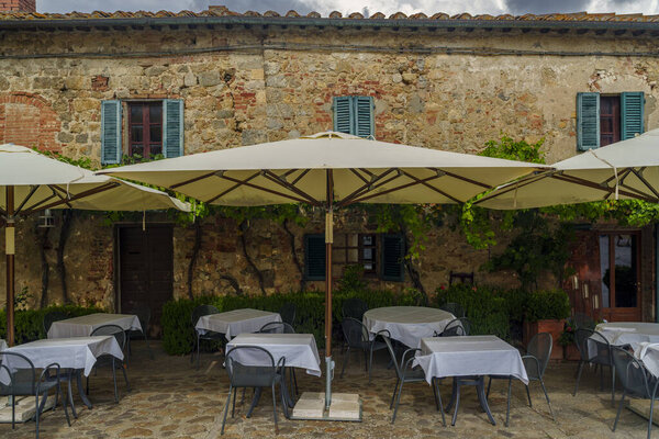 Detail of cafe terrace, cream umbrellas, stone wall, climbing vines, green shutters, cozy Tuscan setting.
