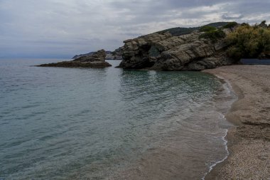 Yumuşak dalgalar, alçak bir kaya kemerinin ve bulutlu gökyüzünün altındaki katmanlı kayalıkların yanında dar çakıllı bir plajı siler..