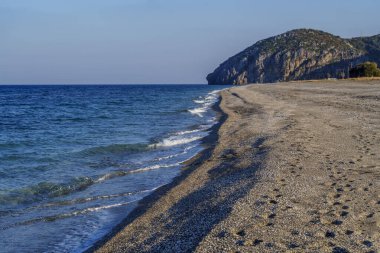 Yumuşak dalgalı boş çakıl taşı plajı, açık mavi deniz ve ufukta kayalık burun..