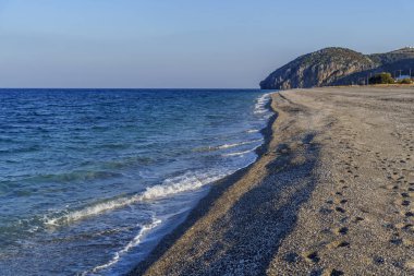 Yumuşak dalgalı boş çakıl taşı plajı, açık mavi deniz ve ufukta kayalık burun..