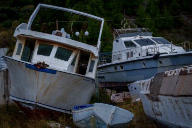 Birkaç yıpranmış tekne, çimenli bir tersanede eğilmiş ve paslanmış, soyulmuş boyaları ve lekeli gövdeleri arkalarındaki yeşil yamaçla çelişiyor..