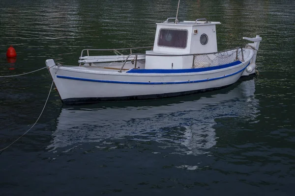 Küçük beyaz ve mavi bir balıkçı teknesi dalgalı liman suyunda sessizce yüzer ve yüzeyde yumuşak bir yansıma oluşturur..