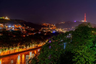 Parlak sokak lambaları sakin nehri yansıtırken, geniş bir yamaç bölgesi ve karanlık çöktükten sonra kırmızı TV kulesi parlıyor..