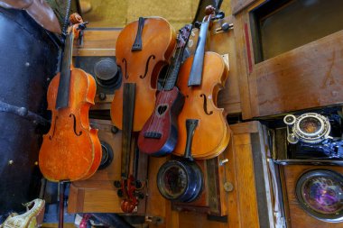 Eski kameralar ve mekanik parçalarla çevrili ahşap raflarda birkaç aşınmış tel enstrüman duruyor..