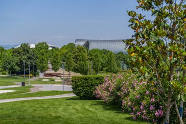 Çimleri, yolları ve pembe çiçekli çalıları olan güneşli şehir parkı uzaktaki gümüş tüp şeklindeki kültür merkezini çerçeveliyor..
