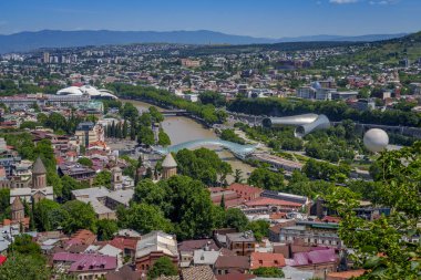 Geniş şehir manzarasında Eski Tiflis çatıları, birden fazla kilise, Barış Köprüsü ile Kura Nehri ve uzak dağlara karşı modern tubular Rike Park konser salonları görülüyor..