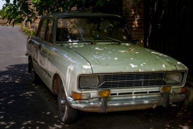 Eski soluk yeşil Sovyet döneminden kalma bir araba, ağaç dallarının altında gölgeli bir sokakta kullanılmadan duruyor.