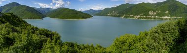 Orman dağları ve parçalı bulutlu gökyüzü ile çevrili sakin mavi rezervuarın geniş panoramik manzarası
