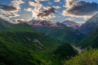 Ilık akşam ışığı yeşil dağ vadisinin üzerindeki bulutları ve uzak kar zirvelerini delip geçiyor.