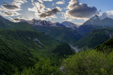 Koyu yeşil vadinin üzerindeki altın gün batımı ışığı ve parlayan bulutların altındaki uzak kar zirveleri.