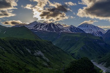 Güneş, derin yeşil dağ yamaçlarının ve uzak karlı zirvelerin üzerinde dramatik bulutların ardında batar.