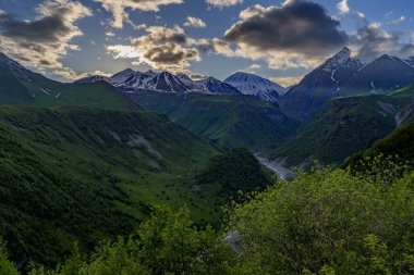 Ilık akşam ışığı yeşil dağ vadisinin üzerindeki bulutları ve uzak kar zirvelerini delip geçiyor.